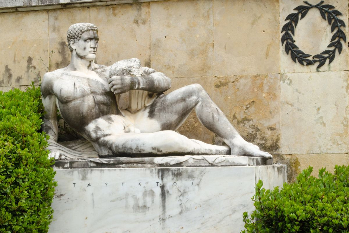 The image shows a statue of a muscular, reclining male figure, representing a warrior, set against a stone wall with a laurel wreath. The statue is partially surrounded by green shrubs.